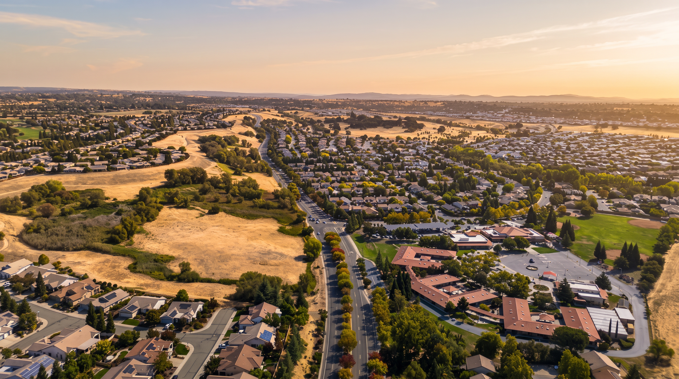 This high-angle aerial perspective captures a sprawling suburban neighborhood characterized by winding residential streets and dense clusters of single-family homes. A prominent multi-lane road cuts through the center of the frame, flanked by mature trees and leading toward a large school complex with athletic fields. The landscape features a mix of developed residential zones and expansive, dry, open fields, highlighting the community's integration with the surrounding natural terrain.