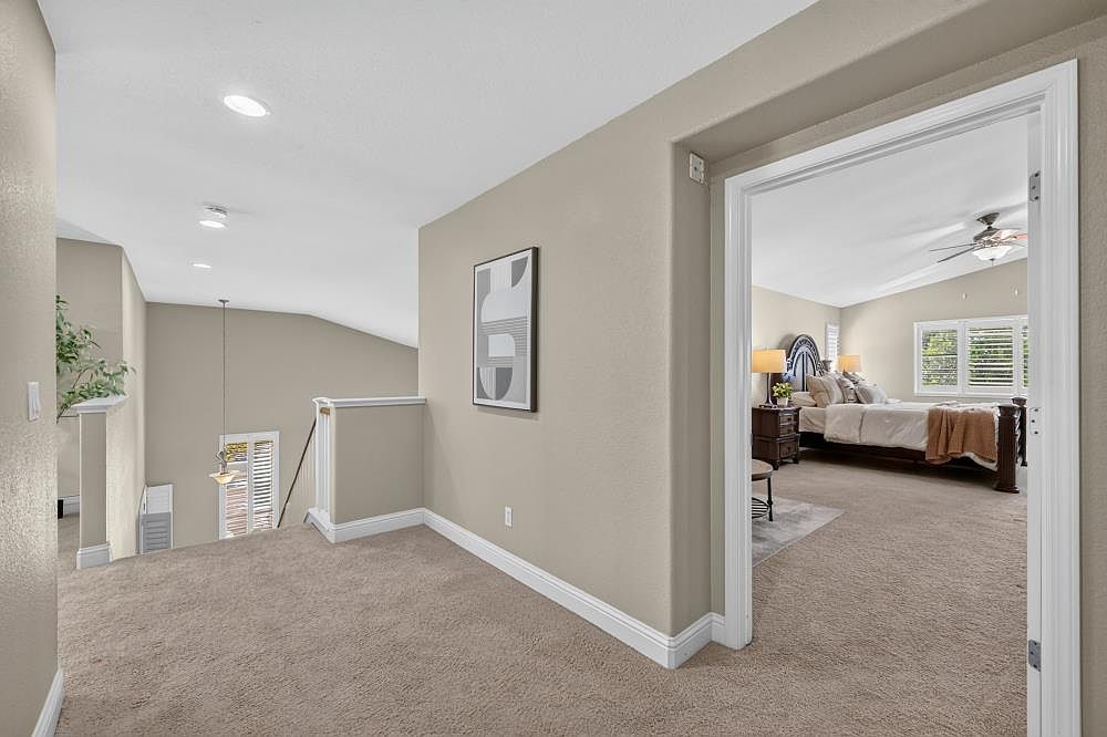 This image captures a carpeted landing area that leads into a primary bedroom through a wide doorway. The neutral-toned walls are accented by a framed piece of abstract art, while a staircase with a white railing descends to the left. The perspective provides a clear transition from the hallway into the bedroom, showcasing a bright, open layout with recessed lighting.