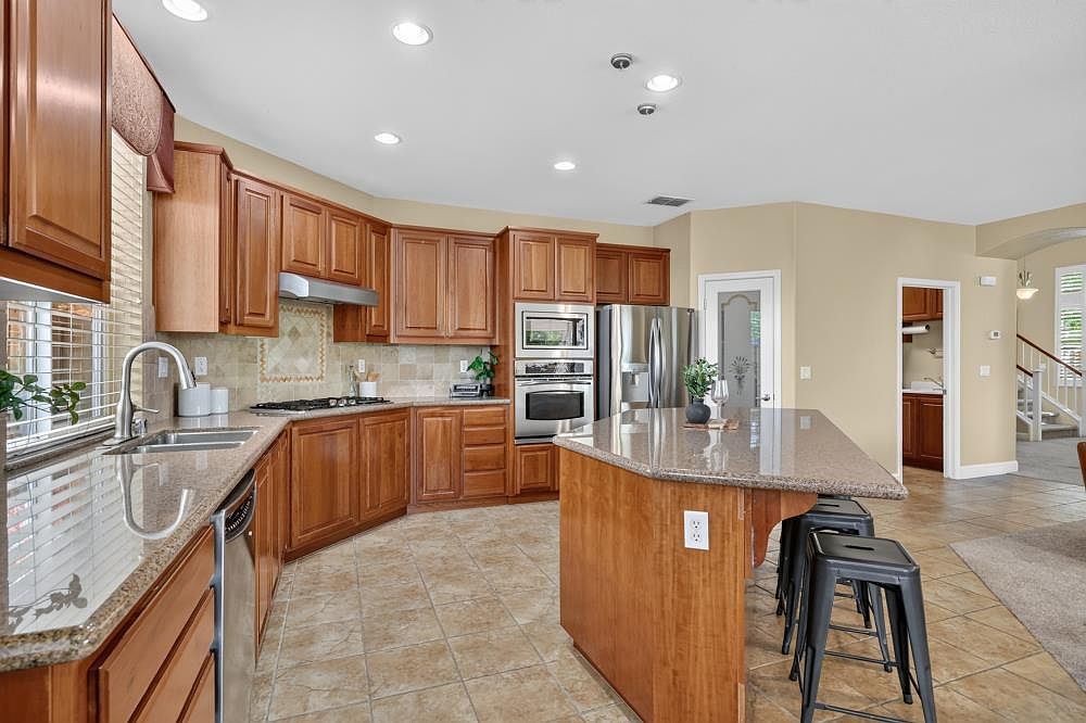 This spacious kitchen features warm-toned wood cabinetry, stone countertops, and a large central island with bar seating. The layout includes stainless steel appliances, a tiled backsplash, and recessed lighting throughout the ceiling. The space flows openly into an adjacent area, creating a functional and inviting atmosphere for cooking and entertaining.