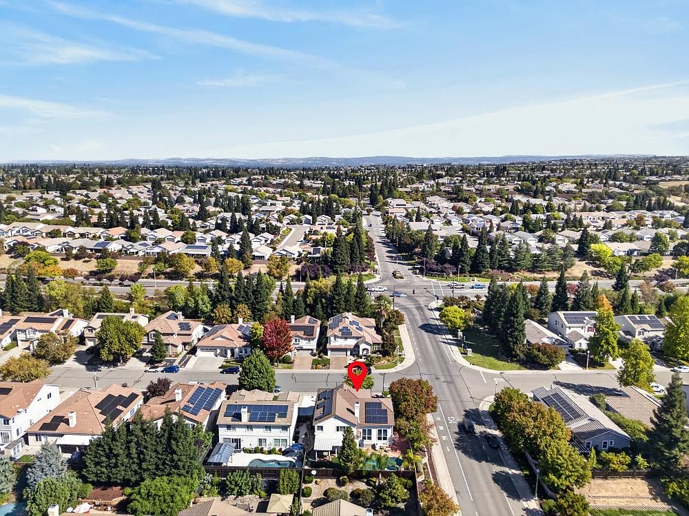 This high-angle aerial view captures a suburban neighborhood on a clear, sunny day, highlighting a residential property marked with a red location pin. The home features a multi-gabled roofline with solar panels, a private backyard with a pool, and a well-maintained landscape. The surrounding area showcases a network of streets, mature trees, and numerous neighboring homes, providing a clear sense of the community layout and proximity to local infrastructure.