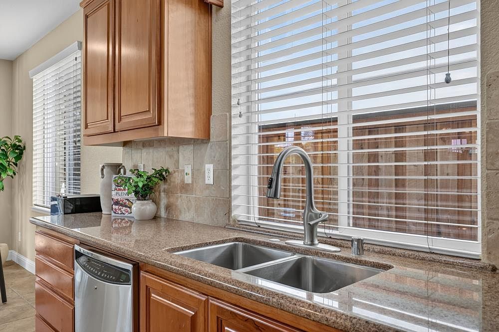This kitchen view highlights a stainless steel double-basin sink with a high-arc faucet set into a speckled stone countertop. Above the counter, wooden cabinets provide storage, while a tiled backsplash adds texture to the wall. A large window with white blinds allows natural light to brighten the workspace, offering a view of the outdoor fencing.