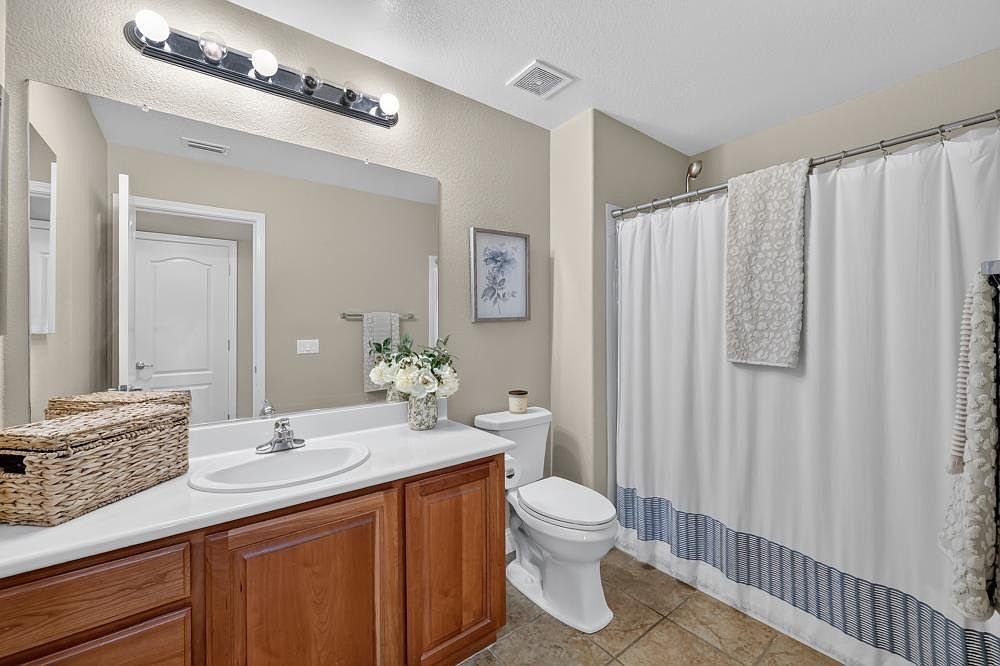 This functional bathroom features a wooden vanity with a white countertop and a single sink, complemented by a large wall-mounted mirror and a multi-bulb light fixture. A white toilet sits beside a shower-tub combination enclosed by a white curtain with a blue-striped pattern. The space is finished with neutral-toned walls and tiled flooring, creating a clean and practical aesthetic.