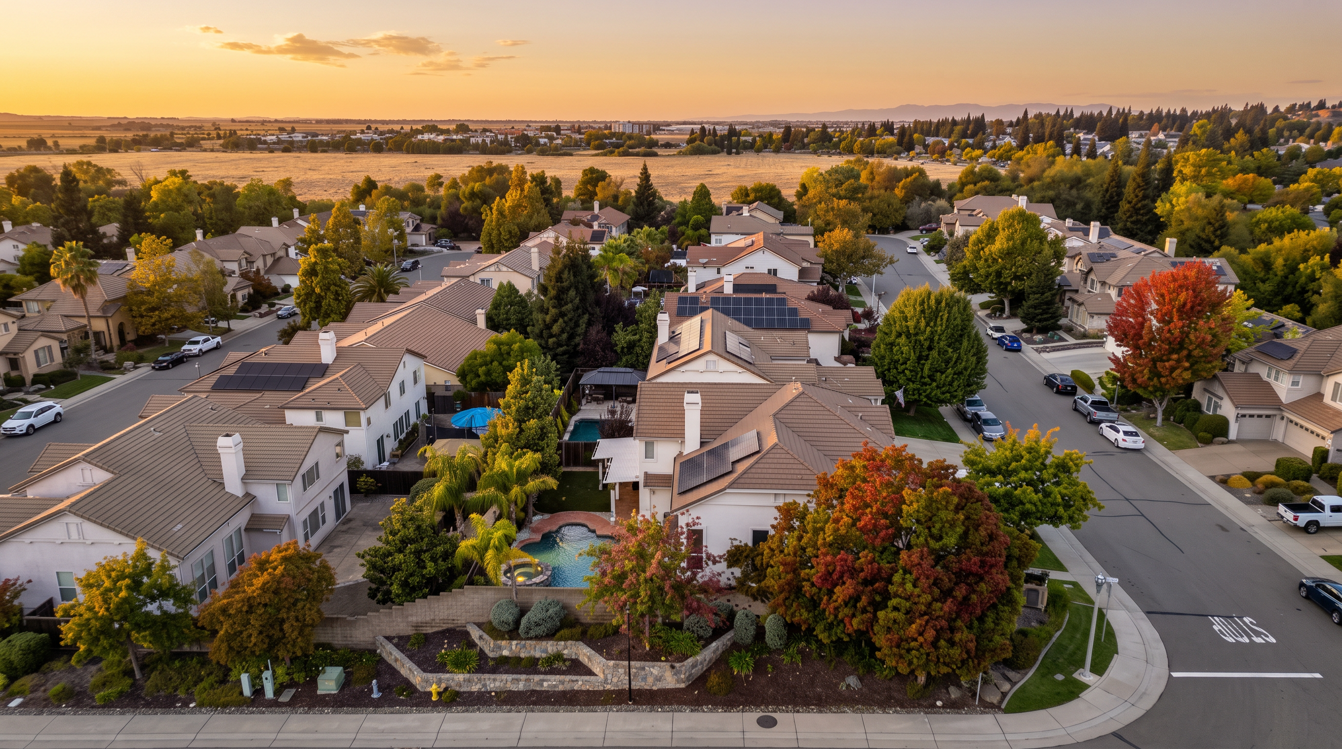 This high-angle aerial view captures a suburban neighborhood featuring several single-family homes with tiled roofs and solar panel installations. The foreground highlights a residence with a private swimming pool, a tiered stone retaining wall, and mature landscaping with colorful autumn foliage. The perspective provides a comprehensive look at the residential layout, street connectivity, and the surrounding open landscape.