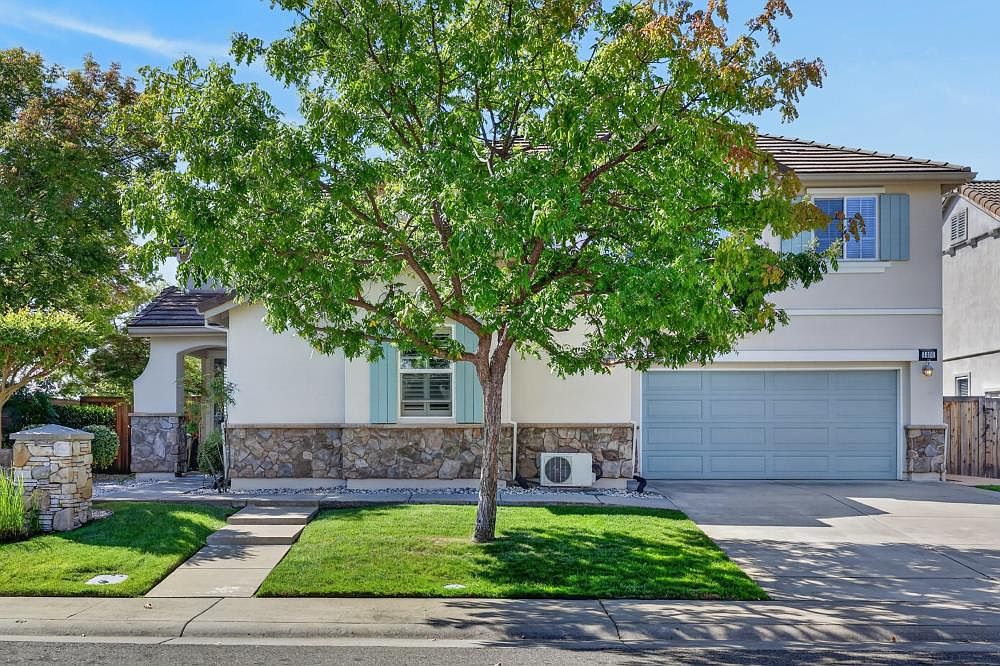This two-story suburban home features a classic facade with a combination of light-colored stucco and stone veneer accents. A mature tree stands prominently in the front yard, providing shade over the manicured lawn and concrete driveway leading to a two-car garage. The home is accented with light blue shutters and a matching garage door, creating a cohesive and inviting curb appeal.