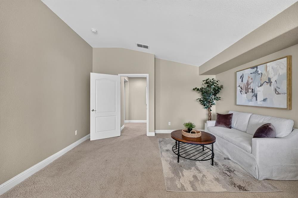 This interior view showcases a cozy living area featuring a light-colored sofa adorned with dark accent pillows and a central oval coffee table. The room is finished with neutral-toned walls, plush carpeting, and a large abstract piece of wall art that adds a contemporary touch. A doorway leads into a connecting hallway, providing a sense of flow and openness to the home's layout.