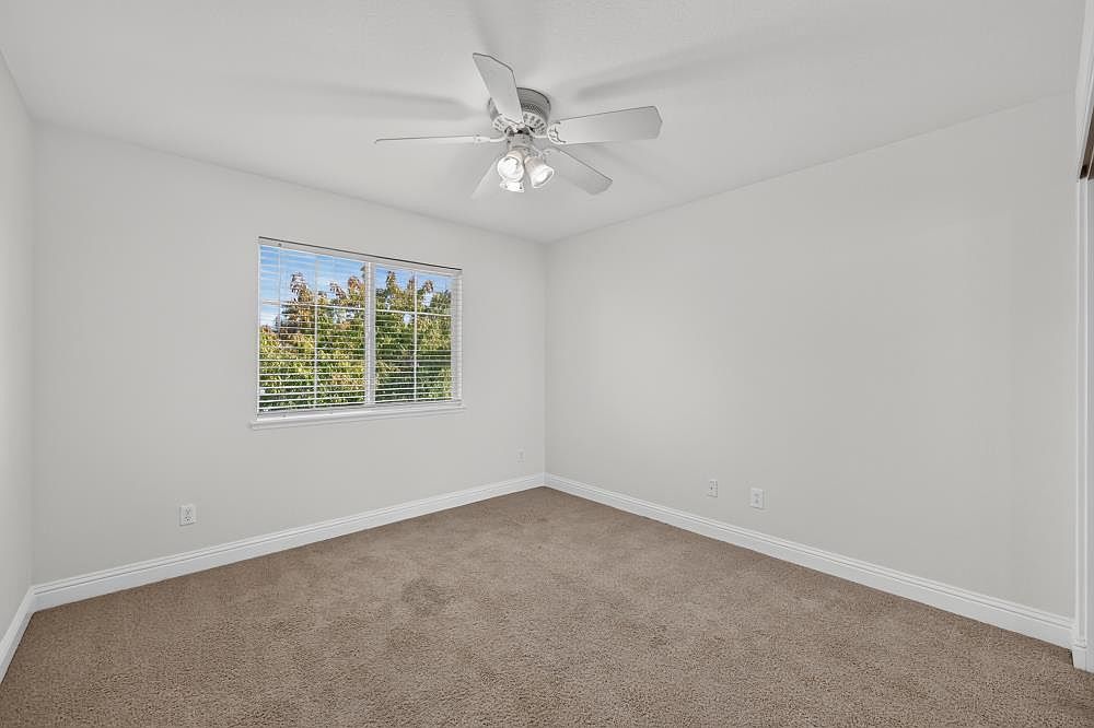 This is a clean, neutral-toned bedroom featuring plush wall-to-wall carpeting and crisp white walls. A ceiling fan with integrated lighting is centered in the room, while a single window with blinds provides natural light and a view of the outdoors. The space is presented as a blank canvas, offering a versatile layout for various furniture arrangements.