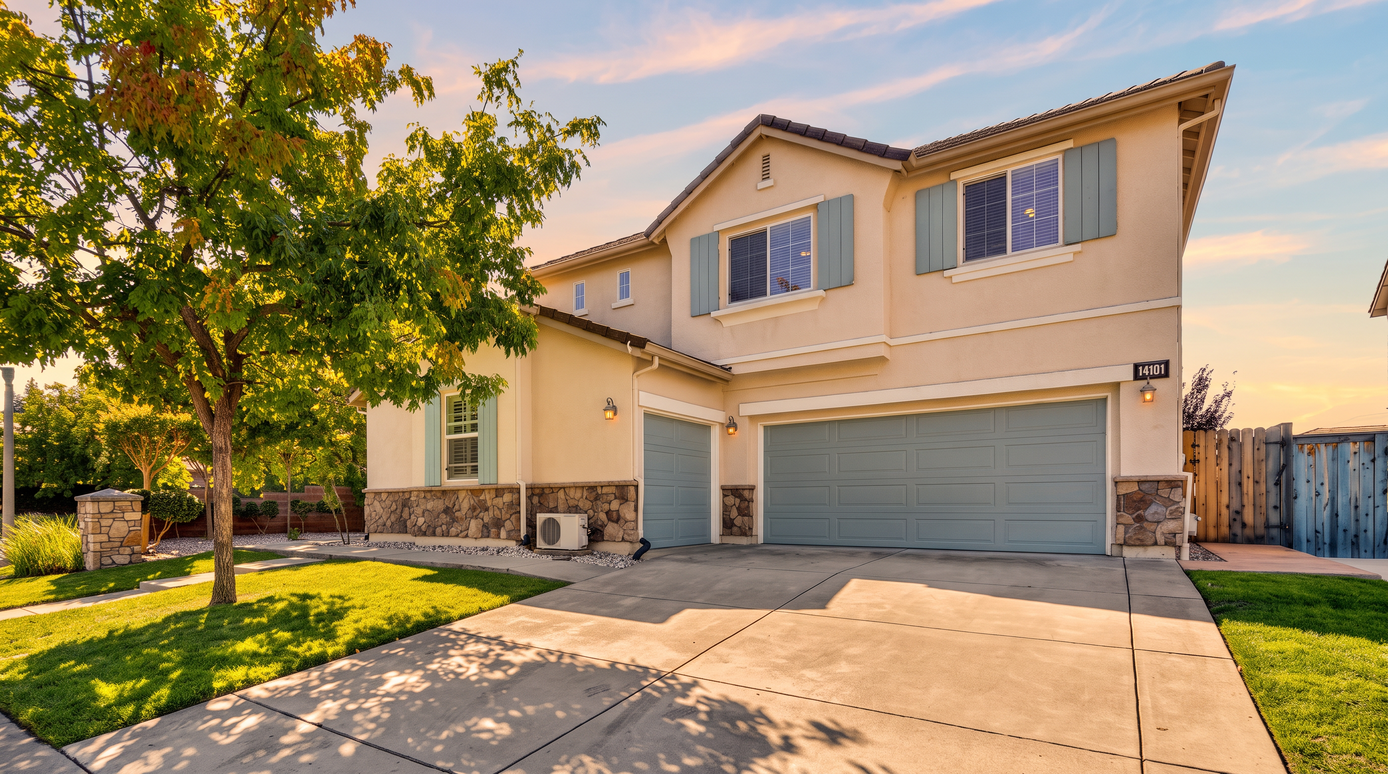This two-story suburban home features a light-colored stucco facade accented with stone veneer along the lower level and blue shutters. The property includes a spacious concrete driveway leading to a three-car garage, complemented by a well-manicured front lawn and a mature tree providing shade. The bright, clear sky and clean landscaping contribute to an inviting and well-maintained curb appeal.