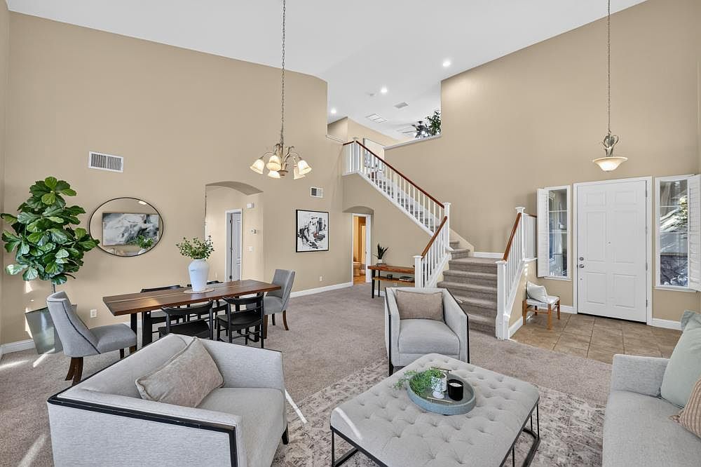 This open-concept living and dining area features high ceilings and a neutral color palette, creating a bright and airy atmosphere. The space includes a formal dining set, a comfortable seating area with a tufted ottoman, and a staircase leading to an upper level. The combination of carpeted floors, elegant light fixtures, and a clear view of the entryway makes this a welcoming and spacious home interior.