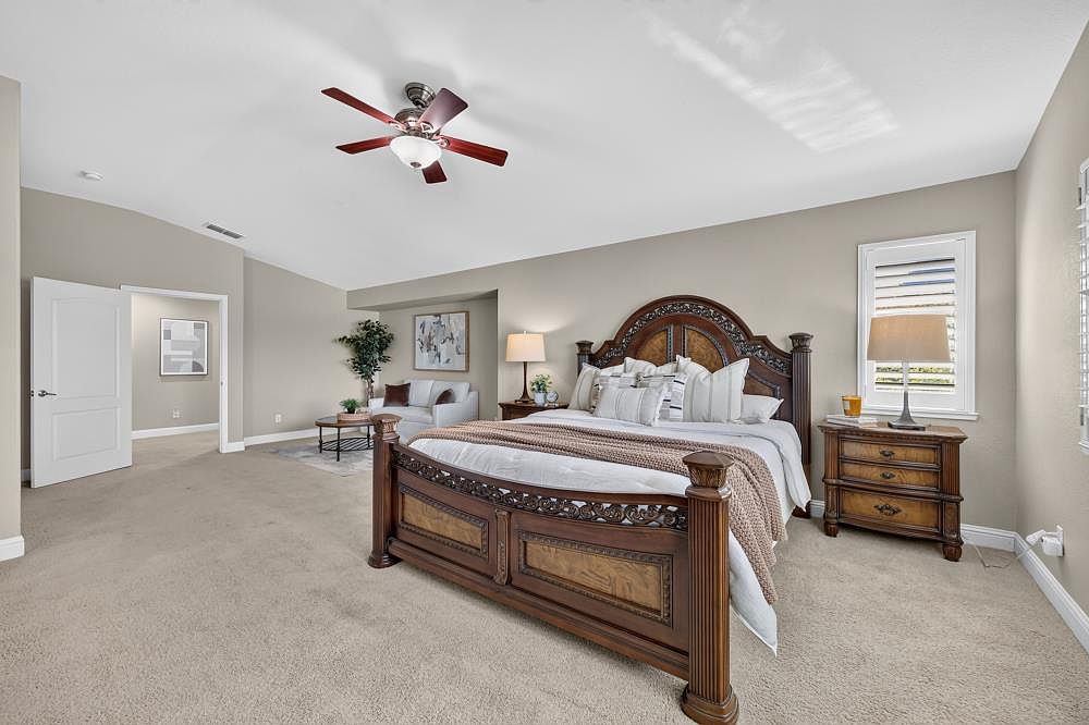 This spacious primary bedroom features a large, ornate wooden bed frame as the central focal point, complemented by matching nightstands and a neutral color palette. A cozy seating area with a sofa and coffee table is tucked into a recessed alcove, creating a versatile living space within the room. The room is illuminated by a ceiling fan with integrated lighting and natural light streaming through a window with plantation shutters.