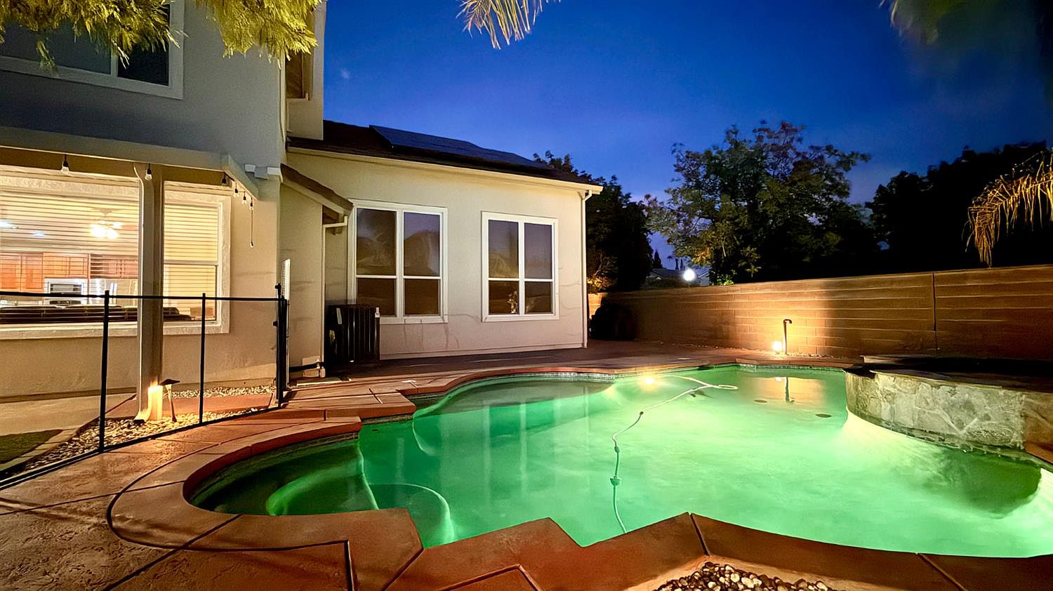 This evening shot captures a backyard oasis featuring a custom-shaped swimming pool with vibrant green underwater lighting. The pool area is bordered by a stamped concrete deck and a black metal safety fence, while the rear of the house displays large windows looking into the interior living space. A tall privacy wall encloses the yard, creating a serene and private atmosphere perfect for nighttime relaxation.