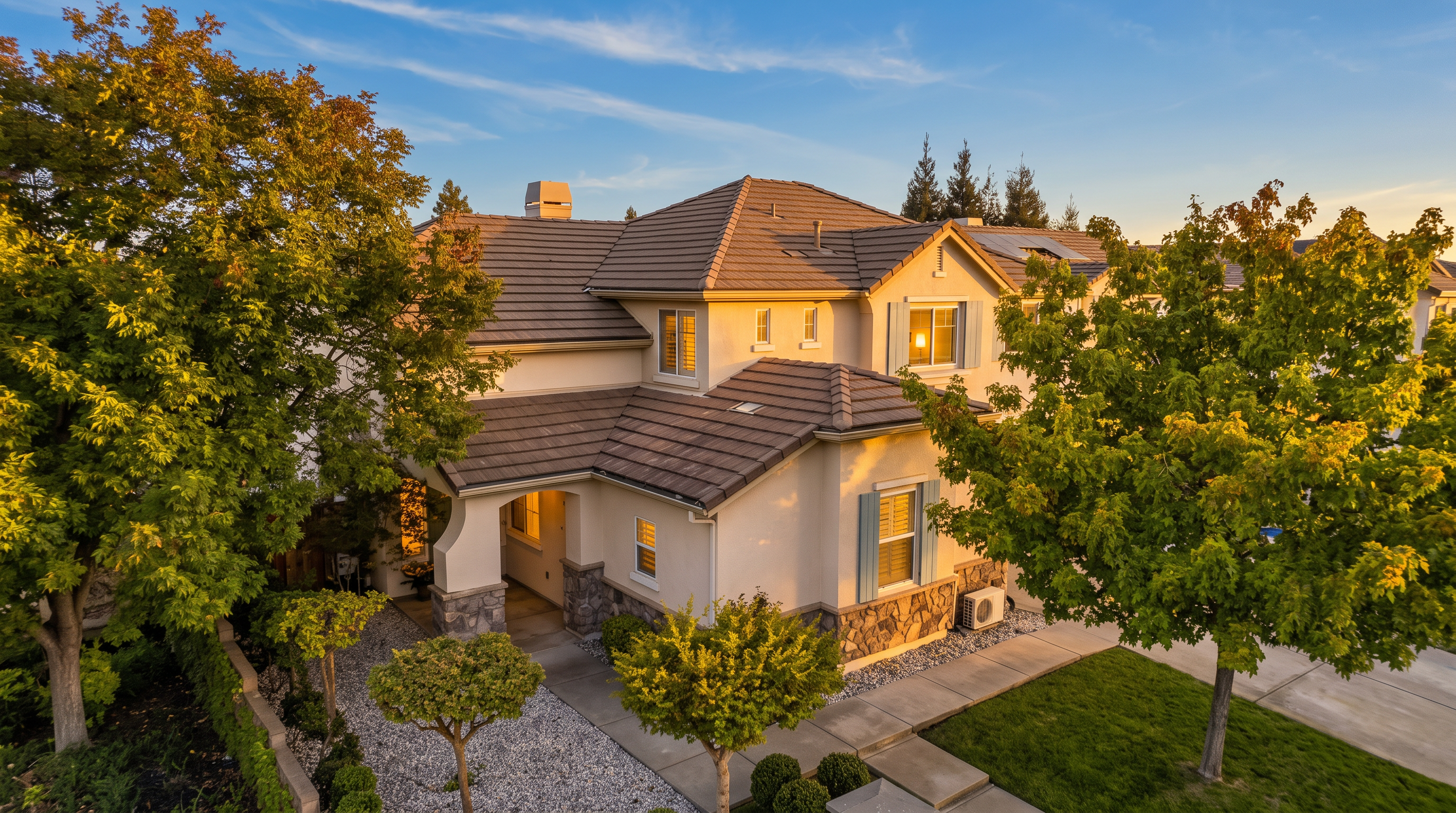 This elevated aerial perspective captures a two-story suburban home with a multi-gabled tile roof and a light-colored stucco facade accented by stone veneer. The front yard features a manicured lawn, a gravel-covered landscaping bed with small, neatly trimmed shrubs, and mature trees that provide shade. A concrete walkway leads to the covered entryway, creating an inviting and well-maintained curb appeal.