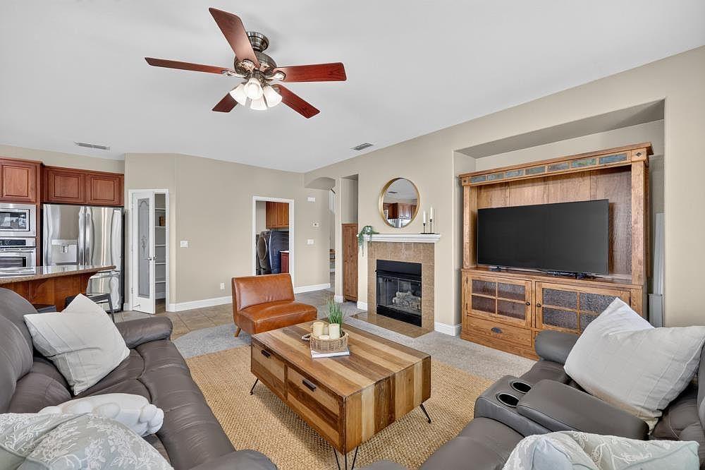 This open-concept living room features a comfortable gray leather sectional sofa and a rustic wood coffee table centered on a textured area rug. A large wooden entertainment center houses a flat-screen television, while a fireplace with a tiled surround and mantel serves as a focal point nearby. The space is bright and inviting, with a ceiling fan overhead and a view leading into the adjacent kitchen area.