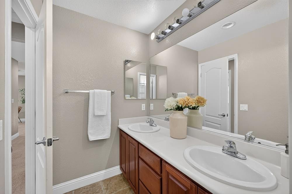 This bathroom features a double-sink vanity with dark wood cabinetry and a light-colored countertop. A large, wall-to-wall mirror reflects the space, which is accented by a decorative vase of flowers and a towel hanging on a wall-mounted rack. The room is finished with neutral-toned walls and tile flooring, creating a clean and functional aesthetic.