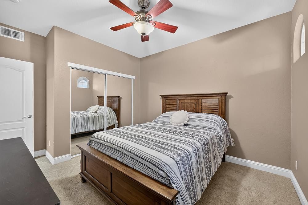 This bedroom features a wooden bed frame with a slatted headboard, dressed in a patterned striped comforter. A large mirrored closet door reflects the room, creating a sense of depth, while a ceiling fan with a light fixture hangs above. The room is finished with neutral-toned walls and carpeted flooring, providing a simple and comfortable living space.