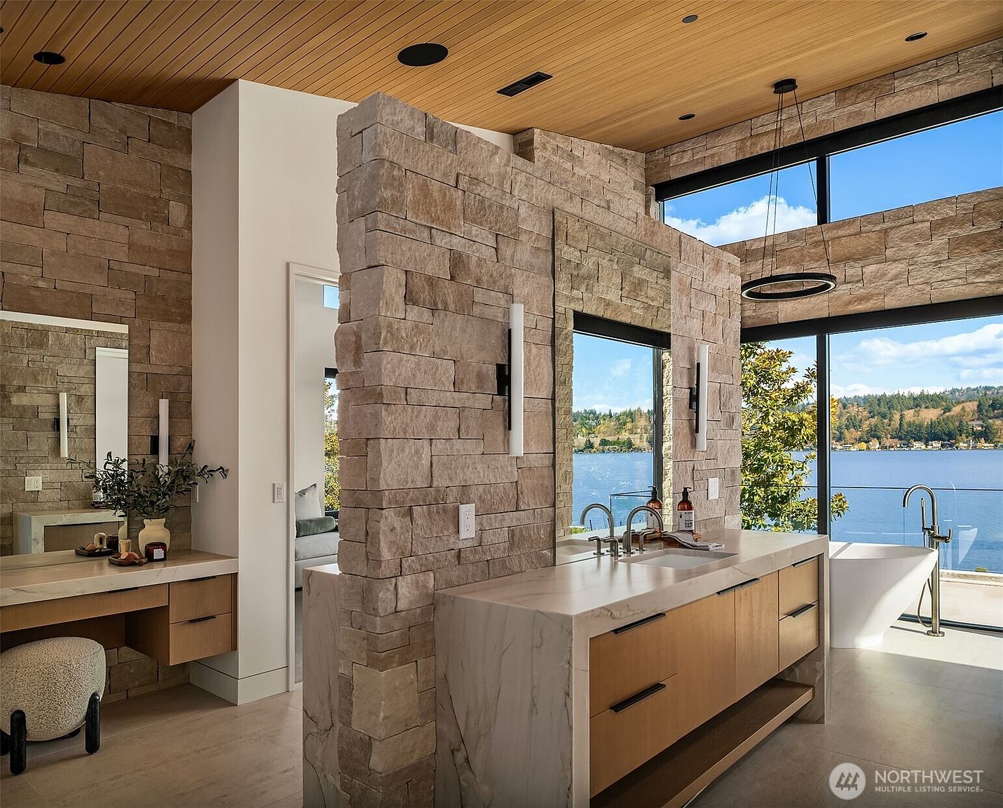 This luxurious primary bathroom features a striking stone-clad partition wall that separates the vanity area from the soaking tub, which overlooks a serene lake view through floor-to-ceiling windows. The space is defined by high-end finishes, including a marble-topped vanity with wood cabinetry, a warm wood-slat ceiling, and modern minimalist lighting. The overall aesthetic is sophisticated and tranquil, blending natural textures with contemporary architectural design.