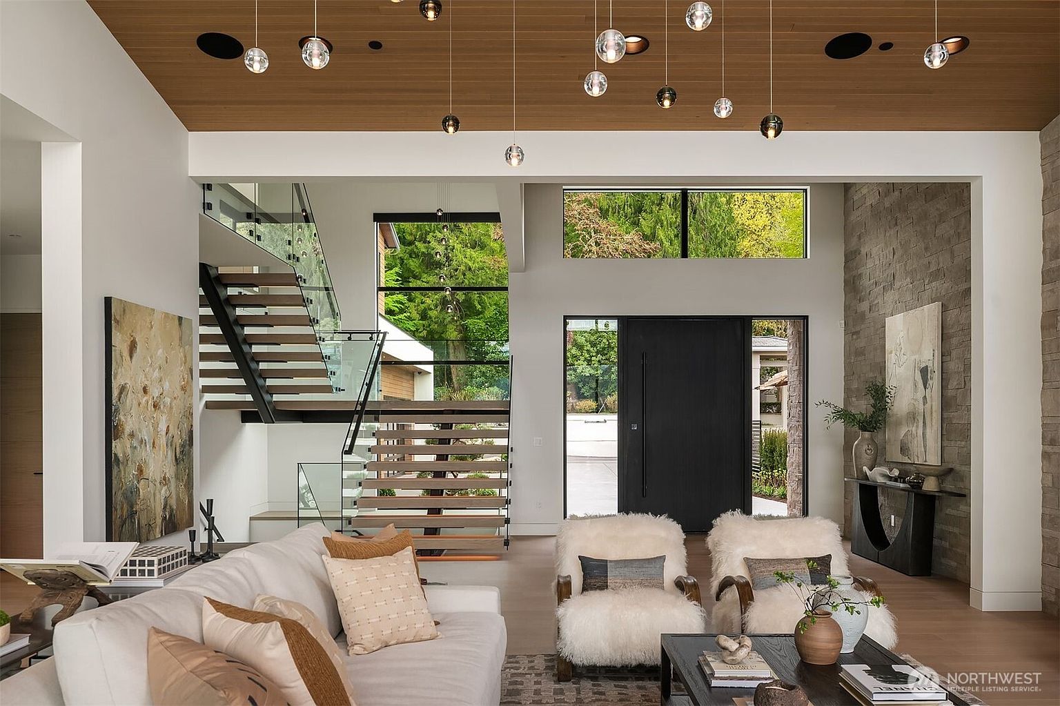 This sophisticated, open-concept living room features a soaring wood-paneled ceiling adorned with a cluster of modern glass pendant lights. The space is anchored by a plush white sofa and two shaggy, fur-covered armchairs, all set against a striking stone accent wall and a dramatic floating staircase with glass railings. Large windows provide a seamless connection to the outdoors, creating an airy, high-end architectural aesthetic.