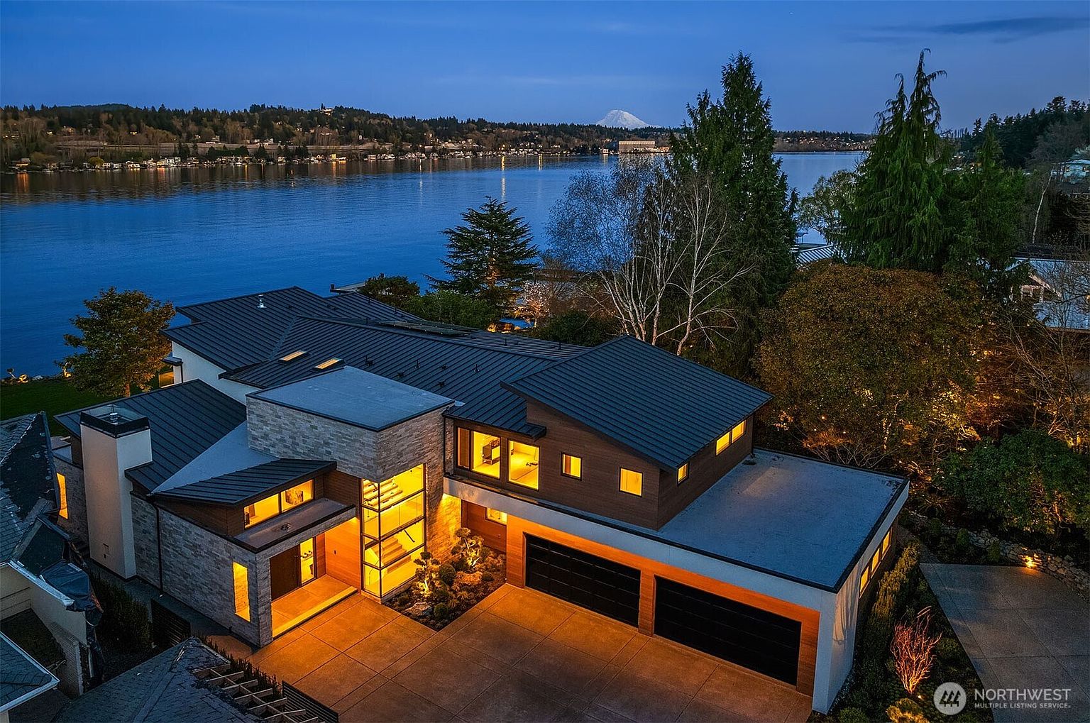 This stunning aerial view captures a modern, multi-level luxury residence at twilight, showcasing its sophisticated architectural design with stone accents and warm, inviting interior lighting. The property features a spacious driveway, a multi-car garage, and a seamless integration with the surrounding landscape, all set against the backdrop of a serene lake and a distant mountain peak. The composition emphasizes the home's contemporary aesthetic and its prime waterfront location.