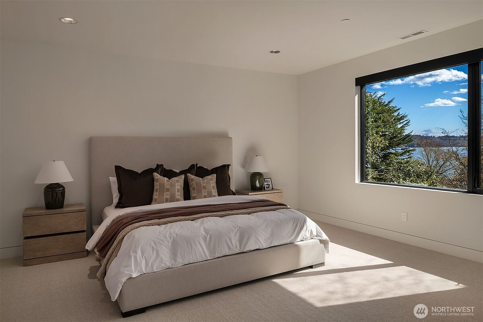 This spacious primary bedroom features a neutral color palette, a plush upholstered bed, and light-toned wood nightstands, creating a serene and modern atmosphere. A large window provides an expansive, picturesque view of the water and distant mountains, flooding the room with natural light. The minimalist design and clean lines emphasize a sense of calm and luxury, perfect for a high-end residential listing.