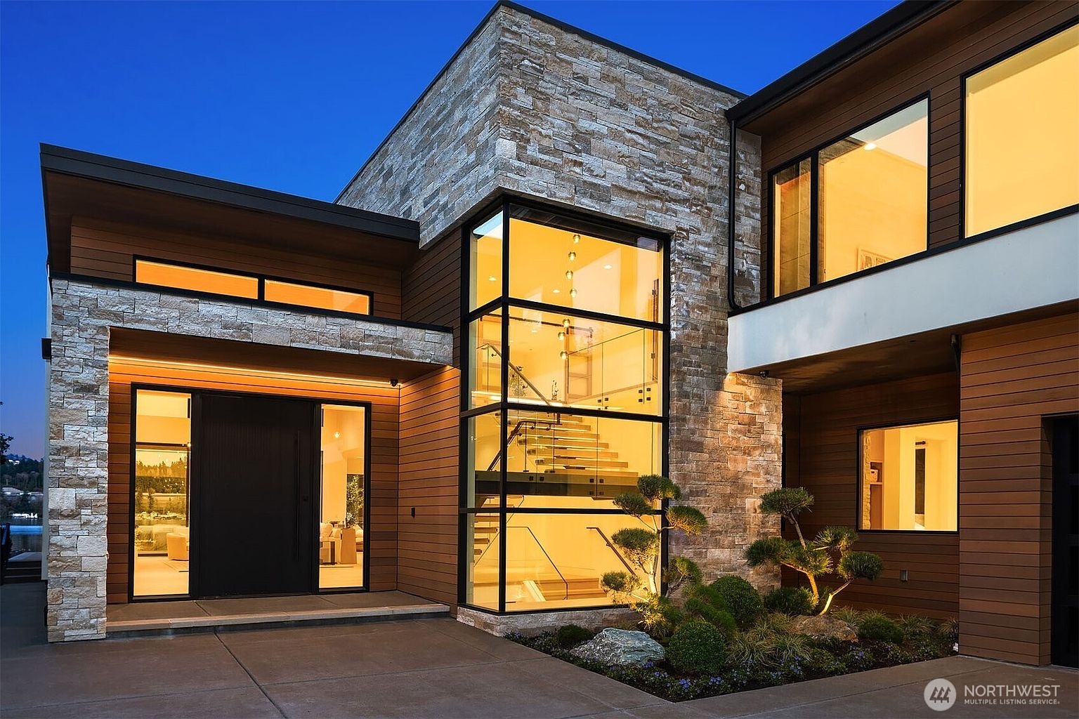 This stunning modern architectural home features a striking blend of natural stone veneer and horizontal wood siding, illuminated by warm, inviting exterior lighting at dusk. A prominent floor-to-ceiling glass tower showcases an elegant interior staircase, serving as a dramatic focal point for the entryway. The scene is completed by a manicured landscape with sculptural trees and a clean, paved driveway, creating a sophisticated and high-end curb appeal.