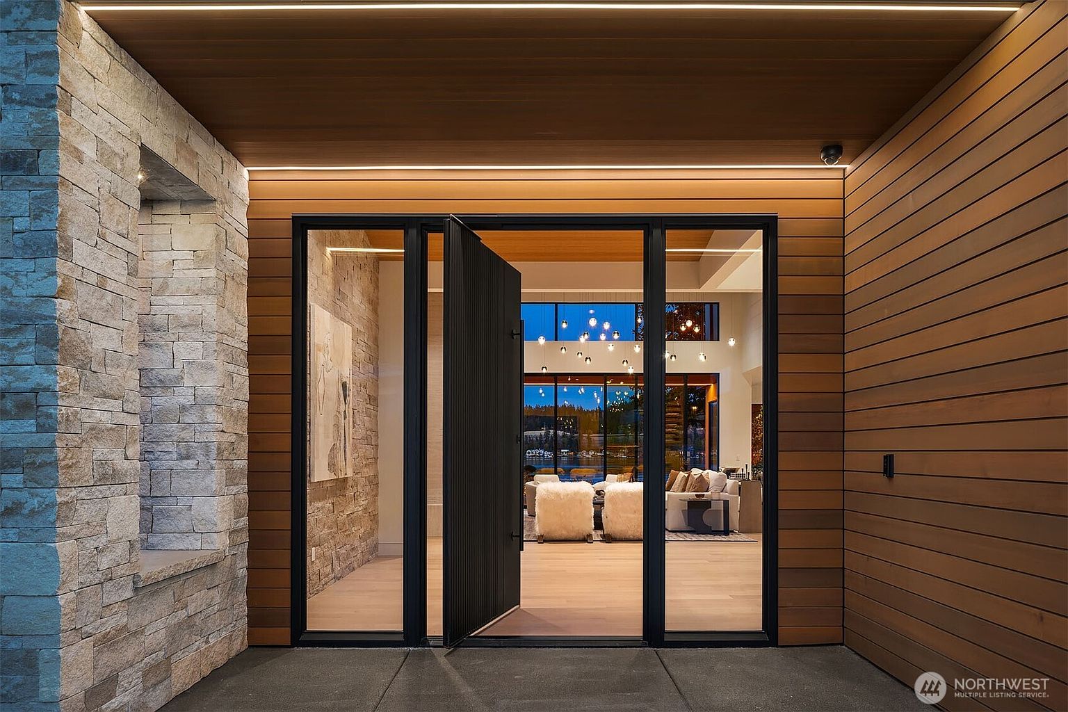 This striking modern entryway features a large, vertical-slatted black pivot door framed by expansive glass panels, creating a seamless transition between the exterior and the interior. The facade combines warm horizontal wood siding with textured stone masonry, accented by integrated linear LED lighting under the soffit. The perspective is a centered, eye-level shot that emphasizes the grand scale and sophisticated architectural design of the home's entrance.