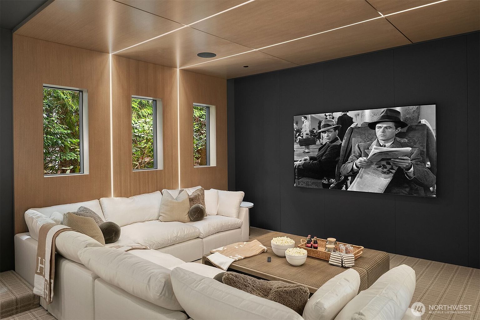 This sophisticated home theater or media room features a large, plush white sectional sofa arranged around a textured coffee table, creating an inviting space for relaxation. The room is accented by warm wood-paneled walls with integrated vertical lighting and a striking matte black feature wall housing a large flat-screen television. The overall aesthetic is modern and luxurious, offering a cozy, cinematic atmosphere perfect for entertainment.
