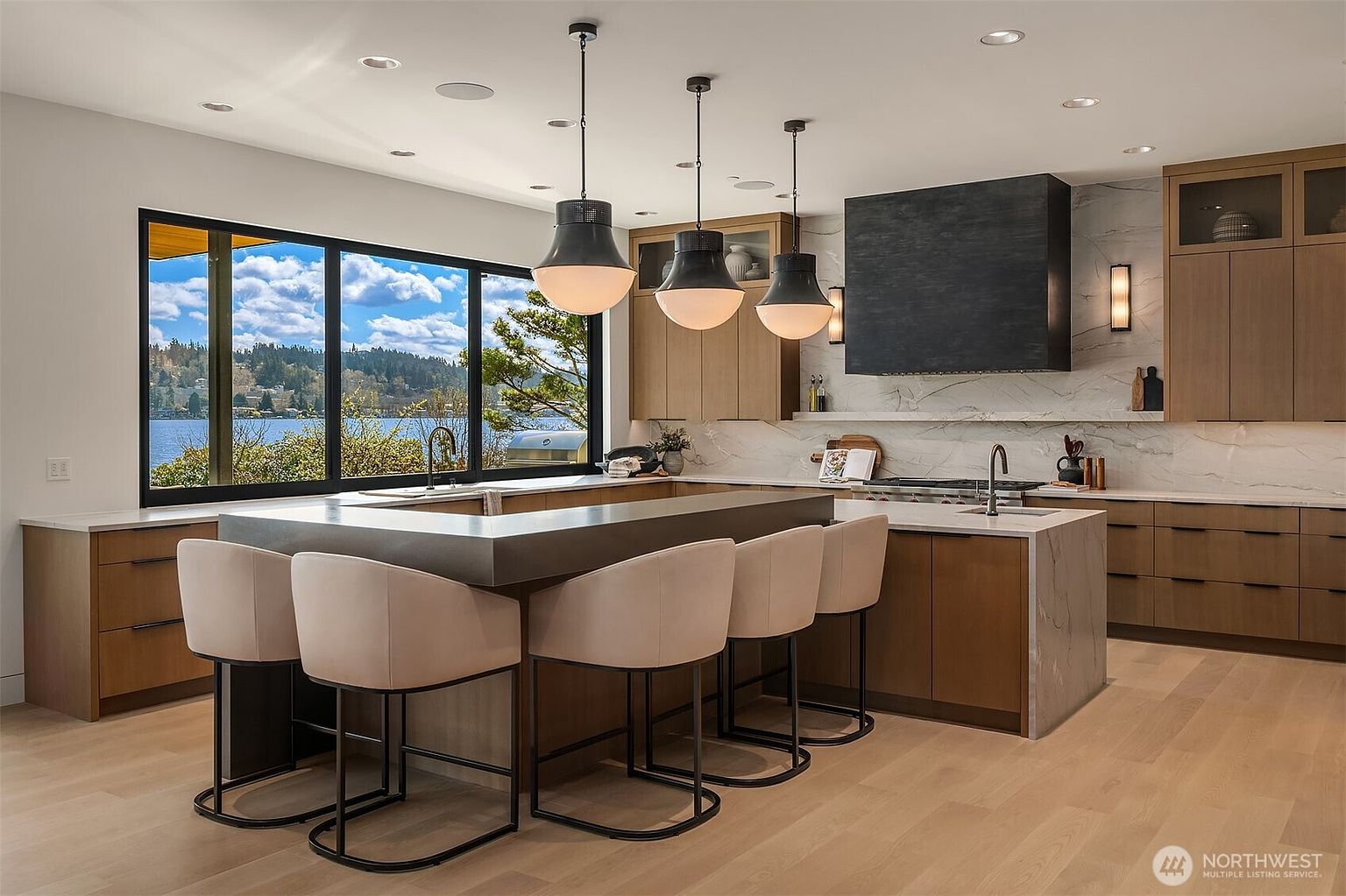 This sophisticated, modern kitchen features a large central island with a dark countertop and integrated seating, complemented by warm wood cabinetry and a striking black range hood. A expansive window provides a stunning, panoramic view of a serene lake and surrounding trees, flooding the space with natural light. The design is elegant and functional, highlighted by stylish pendant lighting and high-end finishes that create a luxurious atmosphere.