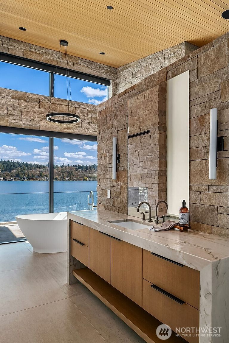 This luxurious primary bathroom features a striking blend of natural stone walls, warm wood cabinetry, and a sleek marble countertop. A freestanding soaking tub is positioned perfectly in front of floor-to-ceiling windows that offer a breathtaking, panoramic view of the lake. The space exudes a modern, spa-like atmosphere with high-end finishes and sophisticated lighting.