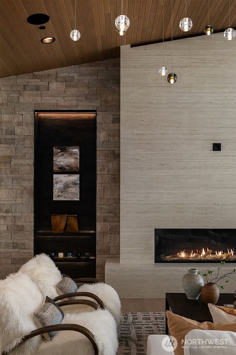 This sophisticated living room features a striking floor-to-ceiling fireplace clad in light-toned, horizontal-veined stone, complemented by an adjacent recessed wall with dark stone cladding and integrated accent lighting. A plush, textured white armchair sits in the foreground, while a collection of modern glass pendant lights hangs from the warm wood-paneled ceiling. The space exudes a luxurious, contemporary aesthetic with a focus on rich textures and high-end architectural details.