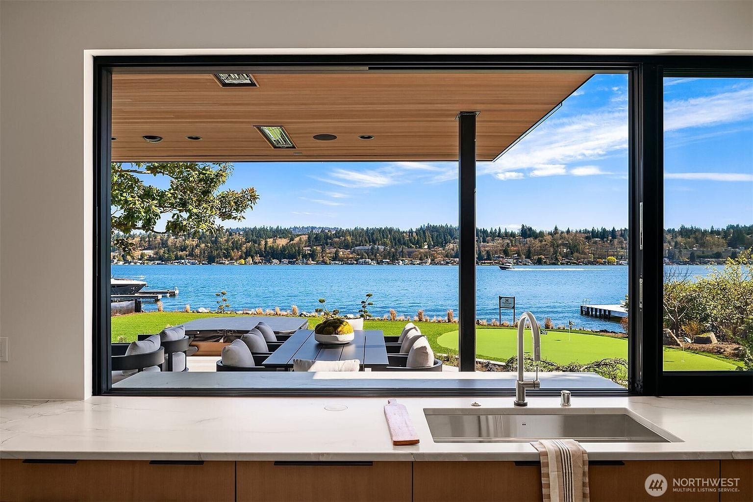 This interior shot captures a sleek, modern kitchen sink area that frames a breathtaking panoramic view of a lake and lush backyard through a large, floor-to-ceiling window. The foreground features a clean marble countertop with a contemporary faucet, while the background reveals a covered outdoor patio with dining furniture and a manicured lawn leading to the water. The composition emphasizes a seamless transition between indoor luxury and the serene natural landscape.