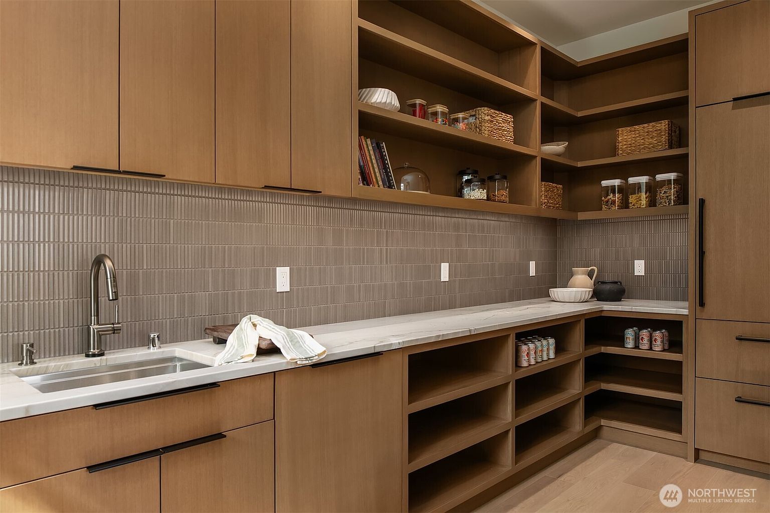 This sophisticated walk-in pantry features warm-toned wood cabinetry, open shelving, and a sleek marble countertop with an integrated sink. The space is accented by a textured, vertical-tile backsplash and modern black hardware, creating a clean, organized, and high-end aesthetic. The perspective is a wide-angle shot that highlights the functional layout and ample storage capacity of this luxury kitchen extension.