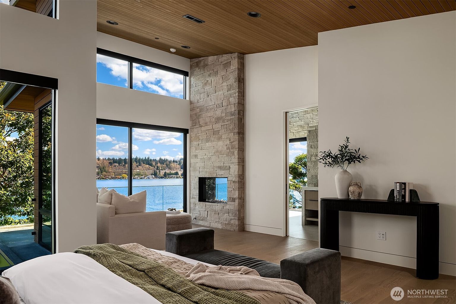 This luxurious primary bedroom features a high, wood-paneled ceiling and a striking floor-to-ceiling stone fireplace that serves as a central focal point. Large sliding glass doors offer expansive, panoramic views of the lake, seamlessly blending indoor comfort with the natural landscape. The room is elegantly furnished with a plush bed, a dark console table, and a minimalist aesthetic that emphasizes light and space.