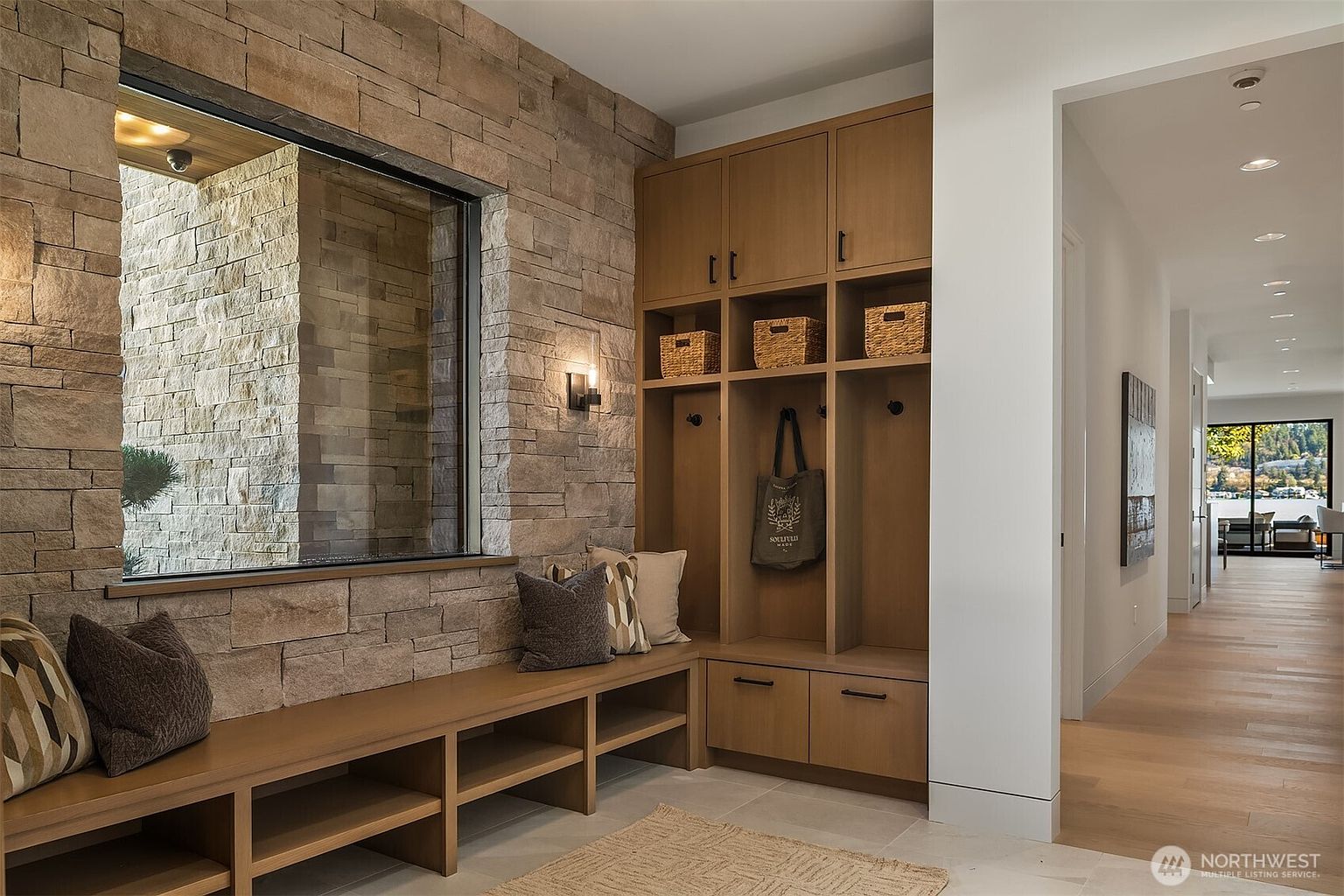 This sophisticated entryway features a custom built-in mudroom bench with integrated storage and a striking stone accent wall that frames a large window. The warm wood tones of the cabinetry and flooring create a welcoming atmosphere, while the open transition into a bright hallway provides a glimpse of the home's expansive layout. The perspective is eye-level, capturing the functional elegance and high-end finishes of this transitional-style mudroom area.