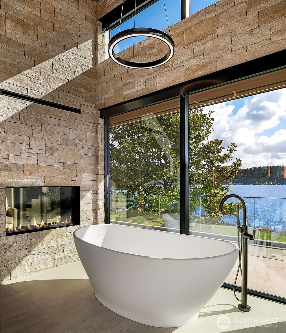 This luxurious primary bathroom features a sleek, freestanding white soaking tub positioned next to floor-to-ceiling windows that offer a stunning, unobstructed view of a lake and lush greenery. A modern linear fireplace is built directly into the textured stone wall, adding warmth and sophistication to the space, while a minimalist circular pendant light hangs above. The combination of natural stone, expansive glass, and high-end fixtures creates a serene, spa-like atmosphere perfect for relaxation.
