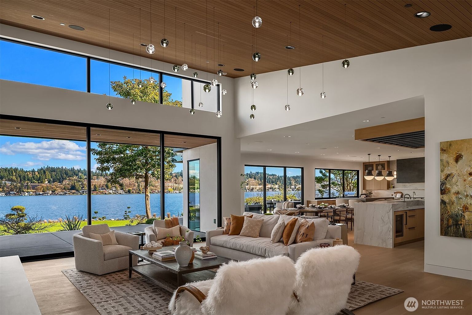This expansive, open-concept living room features a modern aesthetic with high ceilings, warm wood paneling, and floor-to-ceiling windows that frame a stunning waterfront view. The space is furnished with plush, neutral-toned seating, including a large sofa and two textured armchairs, centered around a sleek coffee table. A unique cluster of spherical pendant lights hangs from the ceiling, adding a sophisticated, artistic touch to the bright and airy atmosphere.