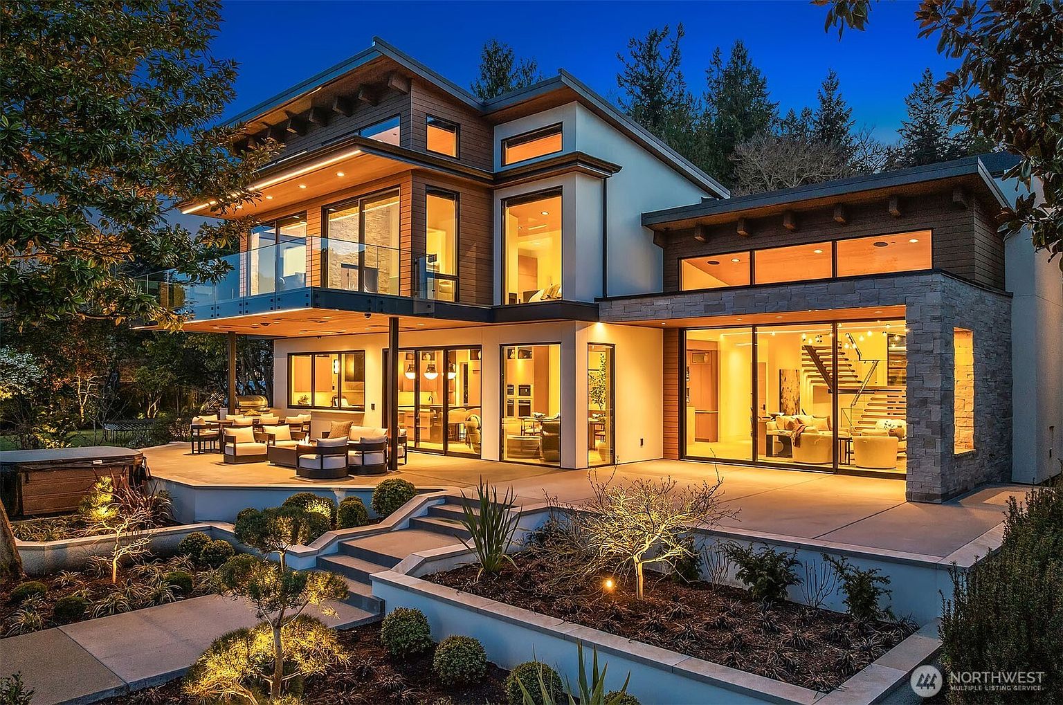 This stunning contemporary residence is captured at twilight, showcasing a multi-level architectural design with expansive glass walls and warm interior lighting. The rear exterior features a spacious concrete patio furnished for outdoor living, complete with a hot tub and tiered landscaping that leads up to the home. The combination of natural wood accents, stone masonry, and modern structural lines creates a sophisticated and inviting atmosphere.
