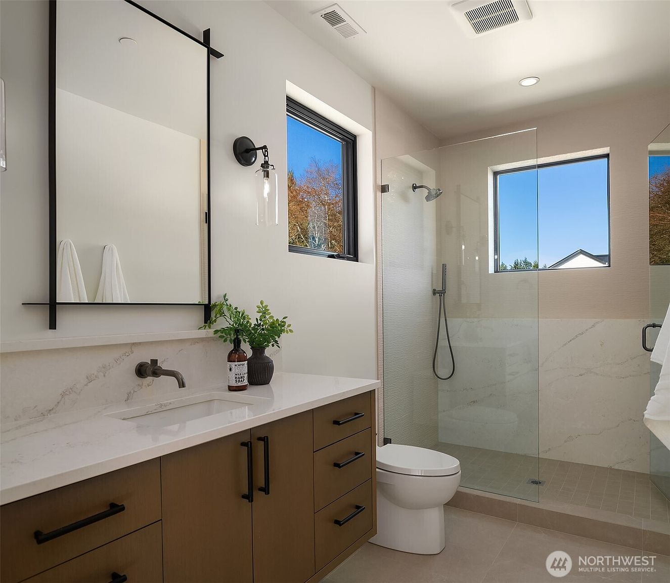 This modern primary bathroom features a sleek vanity with a white countertop, dark wood cabinetry, and a minimalist black-framed mirror. A glass-enclosed walk-in shower with a rainfall showerhead and a window adds a bright, airy feel to the space. The neutral color palette and clean lines create a sophisticated and contemporary atmosphere.