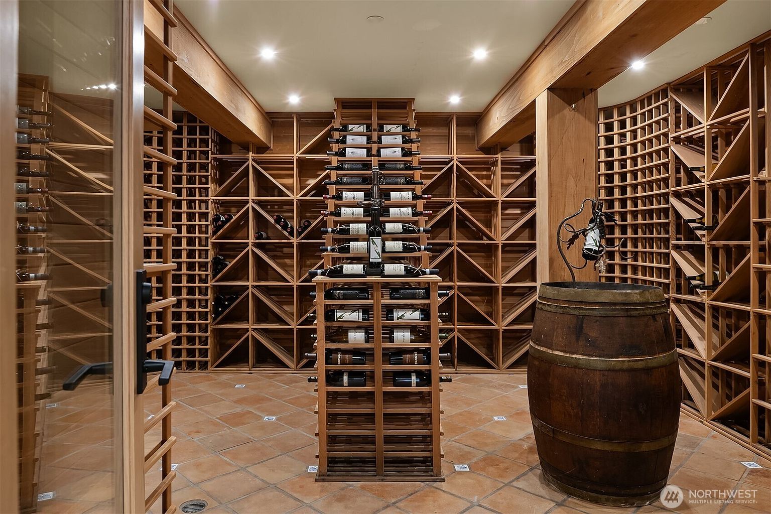 This sophisticated wine cellar features extensive custom wood racking designed to hold a large collection of bottles, with a central display rack showcasing select vintages. A rustic wooden barrel stands in the foreground, adding a classic touch to the space, which is finished with warm terracotta floor tiles and recessed ceiling lighting. The perspective is a centered, eye-level shot that emphasizes the symmetry and organized layout of the cellar.