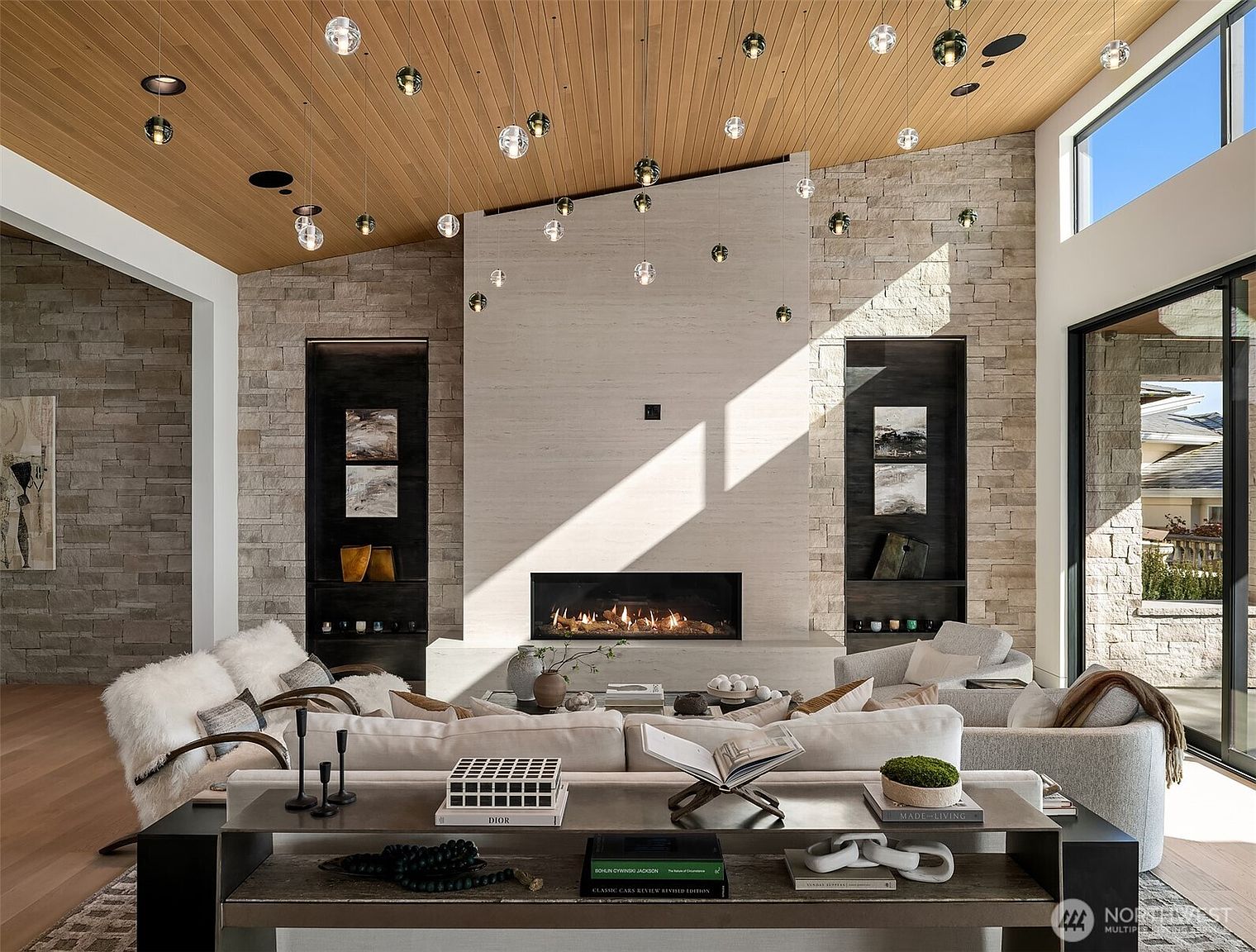 This sophisticated living room features a dramatic floor-to-ceiling stone fireplace as its centerpiece, complemented by a warm wood-paneled vaulted ceiling adorned with a cluster of modern glass pendant lights. The space is furnished with plush, neutral-toned seating and a console table styled with curated decor, creating an inviting and luxurious atmosphere. Large windows and glass doors flood the room with natural light, highlighting the high-end finishes and open-concept design.