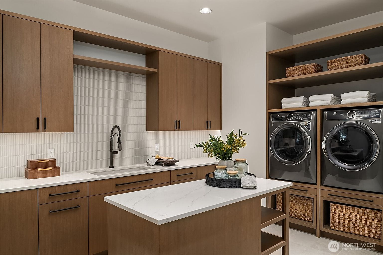 This modern, high-end laundry room features sleek wood-toned cabinetry, a white quartz countertop with an integrated sink, and a central island for folding. The space is equipped with dark-finished, front-loading washer and dryer units, complemented by open shelving and woven storage baskets for a clean, organized aesthetic. The perspective is a straight-on, eye-level shot that highlights the functional layout and sophisticated design of the utility space.