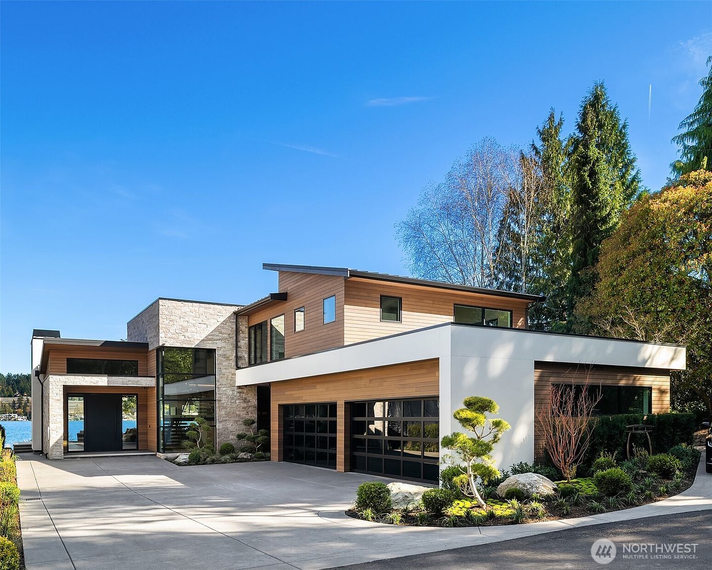 This stunning contemporary residence showcases a sophisticated blend of natural stone, warm wood siding, and crisp white architectural lines. The front facade features a spacious concrete driveway leading to a double garage, complemented by manicured landscaping and a striking glass-enclosed entryway. The perspective captures the home's modern aesthetic and seamless integration with the surrounding environment under a clear blue sky.