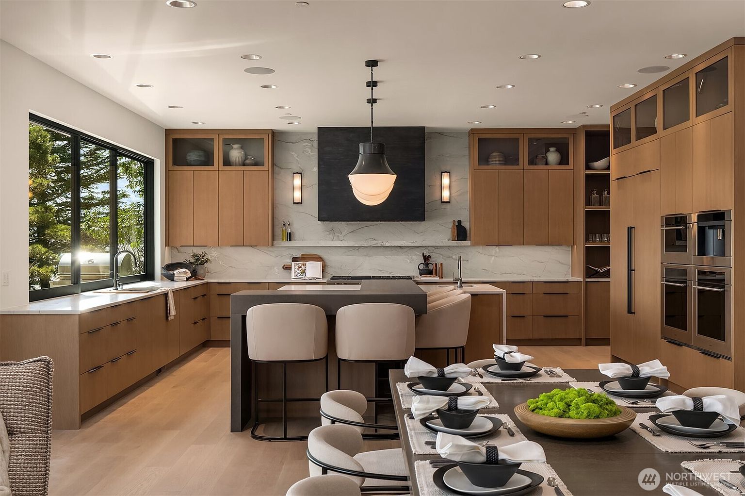 This sophisticated, modern kitchen features warm wood cabinetry, a striking dark charcoal range hood, and a large central island with integrated seating. The space is anchored by a marble-veined backsplash and high-end stainless steel appliances, creating a seamless blend of luxury and functionality. The perspective is a wide, eye-level shot that captures the open-concept flow between the kitchen and the adjacent dining area, emphasizing the bright, airy atmosphere.