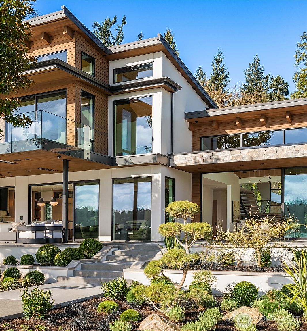 This stunning contemporary residence showcases a sophisticated blend of natural wood siding, crisp white stucco, and expansive floor-to-ceiling glass walls. The multi-level design features a sleek glass-railed balcony and a seamless transition to a manicured landscape with tiered concrete steps and ornamental plantings. The perspective captures the architectural harmony between the modern structure and the lush, serene outdoor environment.
