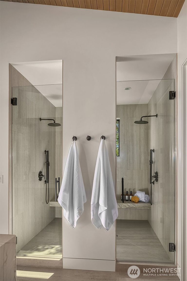 This luxurious primary bathroom features a symmetrical dual-shower design with glass enclosures and light-toned stone tile walls. Each shower stall includes a rainfall showerhead, a handheld sprayer, and a built-in stone bench, while two white towels hang neatly on hooks between the units. The space exudes a clean, modern, and spa-like atmosphere with high-end finishes.