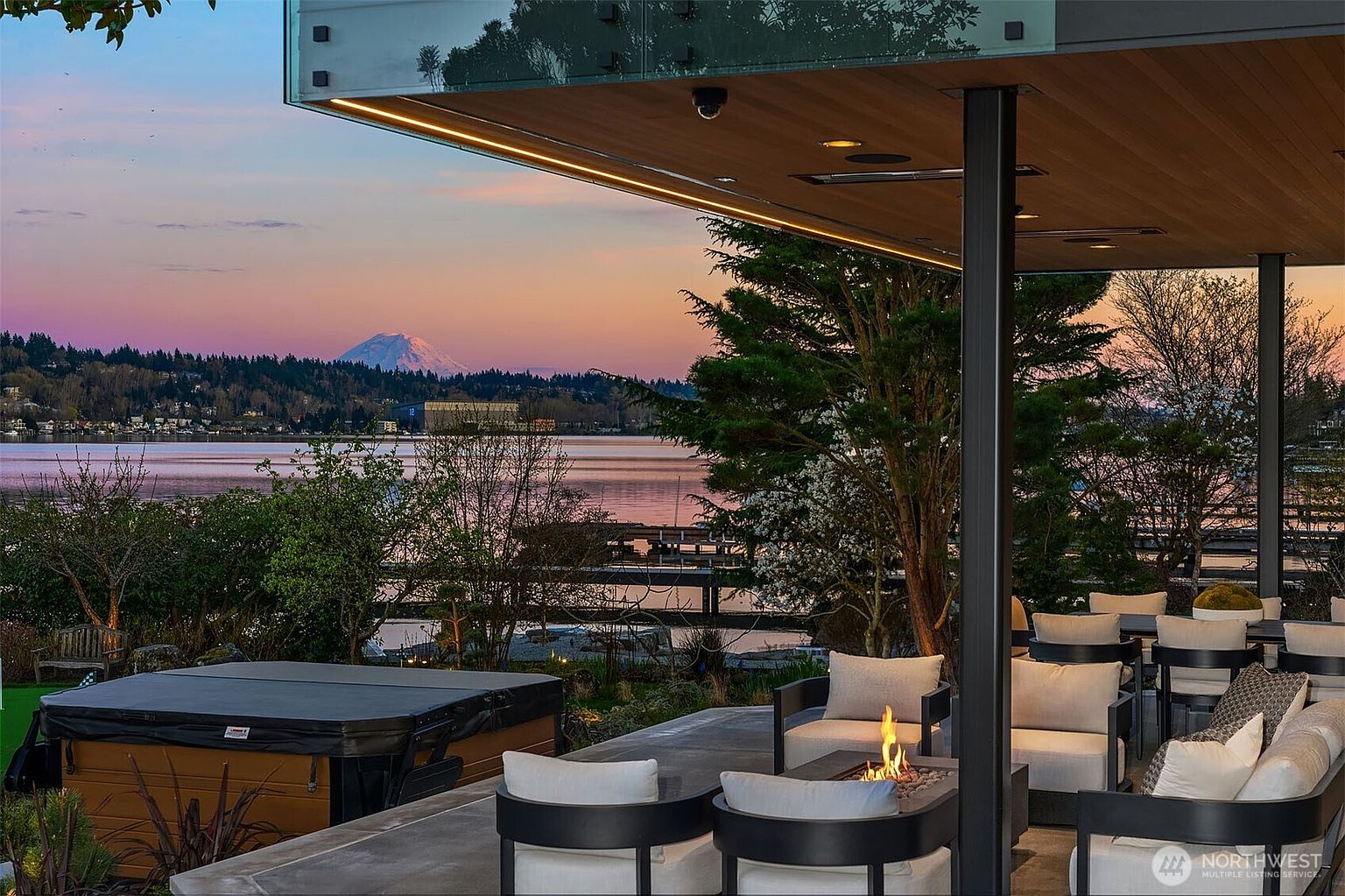This stunning covered patio offers a luxurious outdoor living space with a built-in fire pit, comfortable lounge seating, and a hot tub, all overlooking a serene lake with a majestic mountain view in the distance. The modern architectural design features clean lines and a seamless transition between indoor and outdoor living, perfect for sunset relaxation. The perspective captures the inviting atmosphere of the patio while highlighting the breathtaking natural scenery beyond.