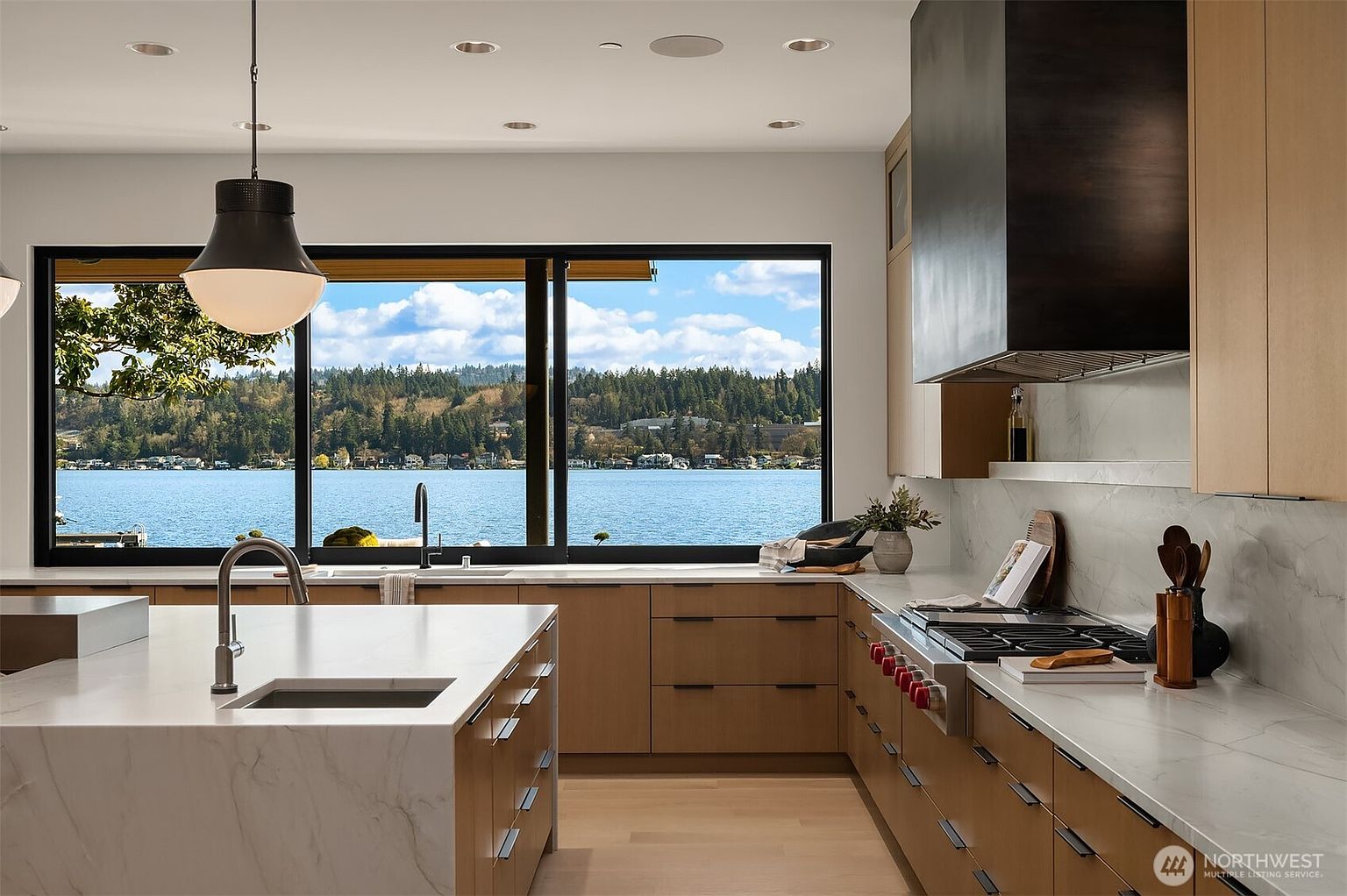 This modern, high-end kitchen features sleek light wood cabinetry, a large waterfall-edge marble island, and a professional-grade range with a custom dark hood. A expansive window provides a stunning, unobstructed view of the lake and surrounding forest, flooding the space with natural light. The design is minimalist and sophisticated, emphasizing clean lines and premium materials.