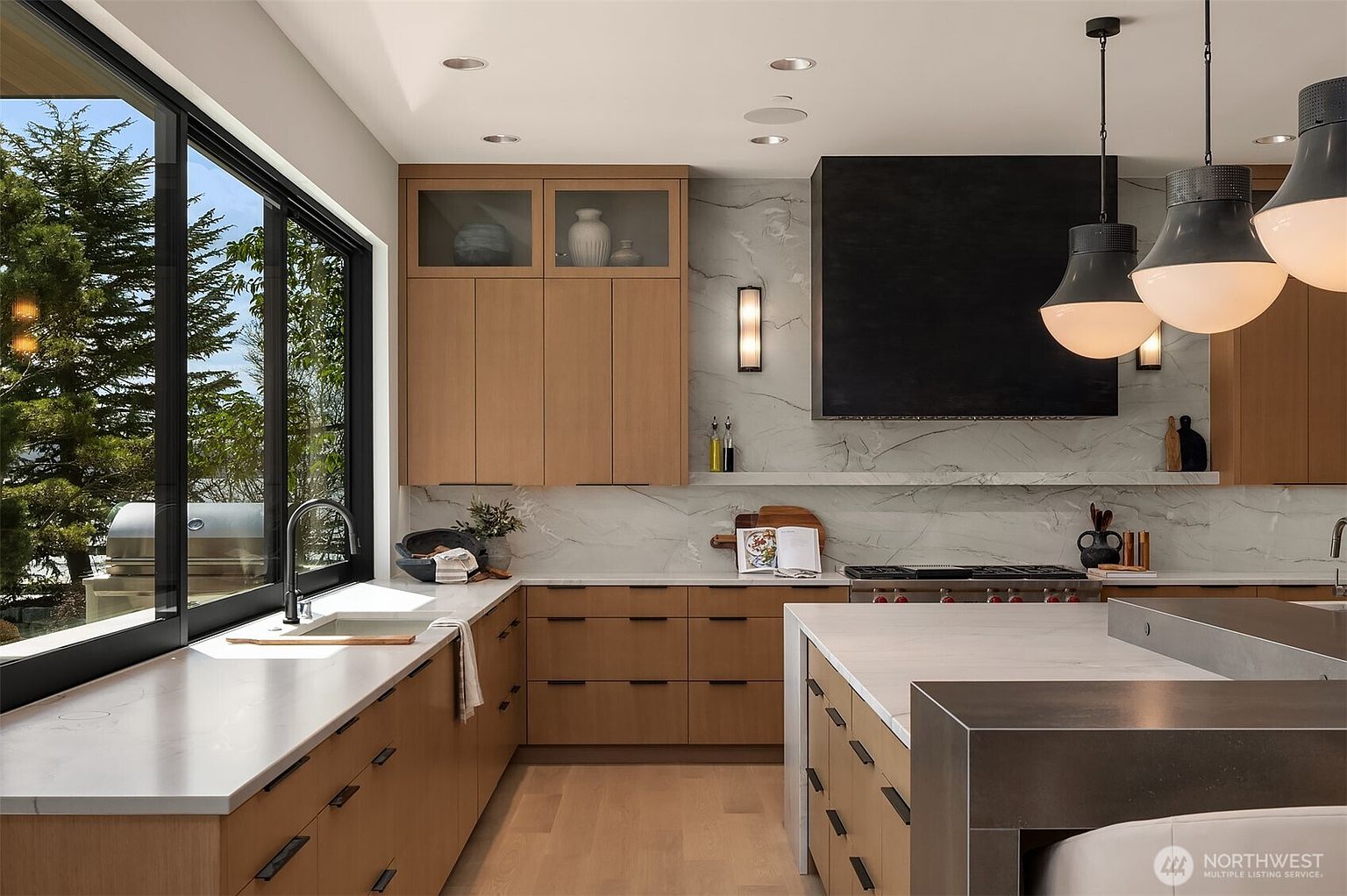 This sophisticated modern kitchen features warm wood cabinetry paired with sleek white marble countertops and a striking black range hood. A large central island provides additional workspace and seating, while expansive windows offer a bright, airy view of the outdoors. The space is accented by contemporary pendant lighting and minimalist hardware, creating a luxurious and functional culinary environment.