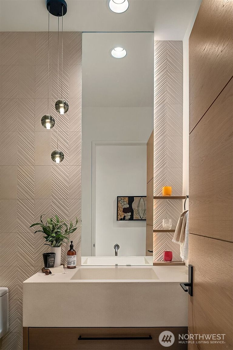 This elegant powder room features a sophisticated, modern aesthetic with textured chevron-patterned wall tiles and a sleek, floating vanity. A tall, frameless mirror reflects the space, complemented by three minimalist glass pendant lights and integrated shelving for decor. The warm wood tones and neutral color palette create a serene and high-end atmosphere.