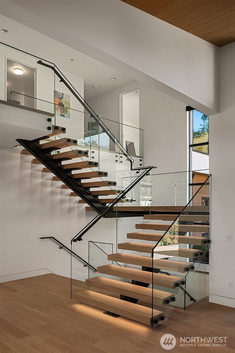 This striking interior space features a modern, open-riser staircase with light wood treads, black metal stringers, and sleek glass railings. The design emphasizes architectural transparency and clean lines, with integrated under-stair lighting that adds a warm, sophisticated glow to the hardwood floors. The perspective captures the dramatic geometry of the staircase as it ascends toward an upper landing, showcasing a contemporary and high-end aesthetic.
