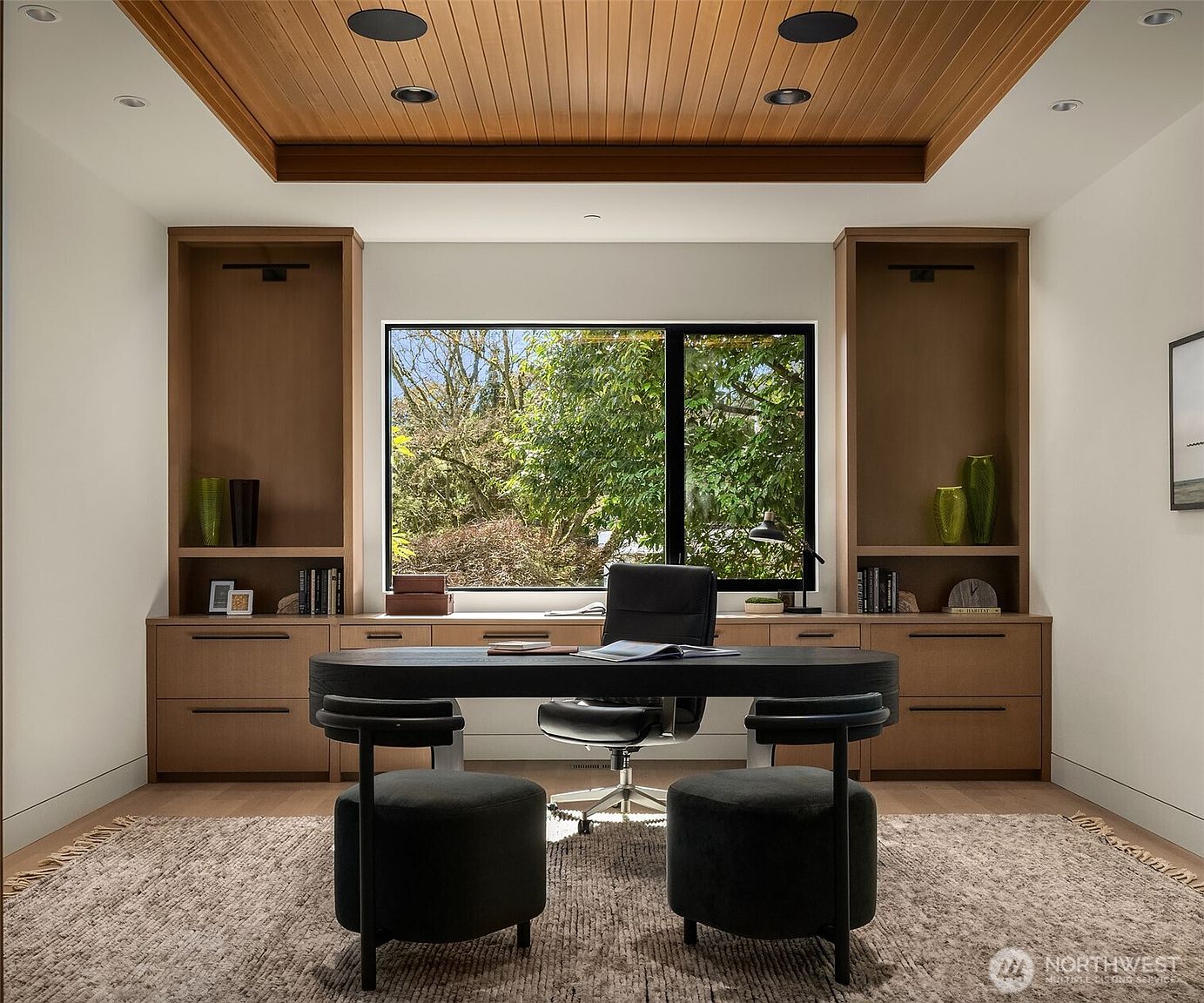 This sophisticated home office features a sleek, dark-toned desk centered in front of a large window that provides a serene view of lush greenery. The room is anchored by custom built-in wooden cabinetry and shelving on either side, complemented by a warm, wood-paneled tray ceiling with recessed lighting. A textured area rug and two modern, dark-upholstered stools complete the professional yet inviting aesthetic.