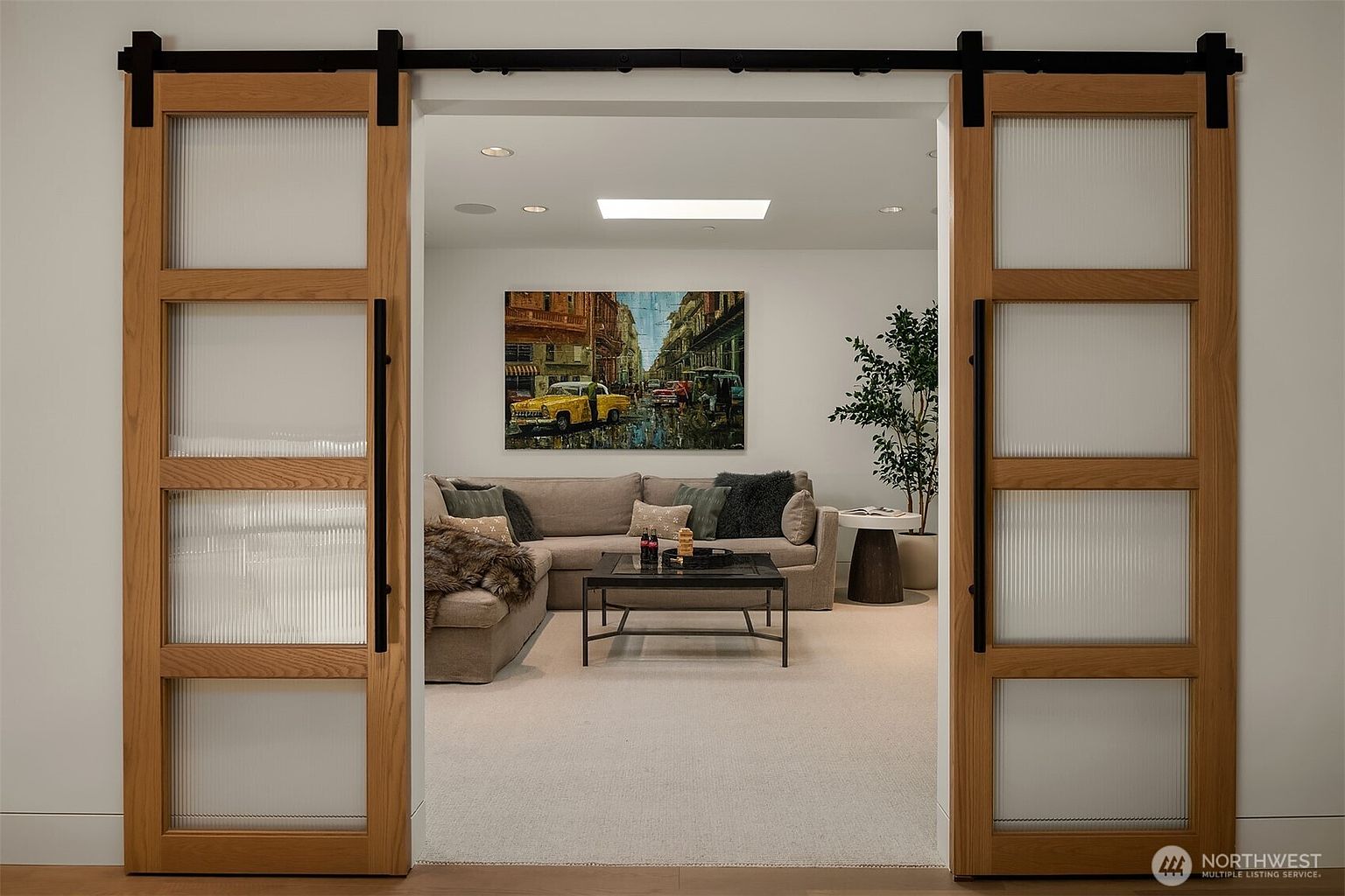 This interior shot is framed through a set of elegant, light-wood sliding barn doors with frosted glass panels, creating a sense of depth and transition. Beyond the doors lies a cozy, well-appointed living area featuring a plush sectional sofa, a central coffee table, and a vibrant cityscape painting on the wall. The space is illuminated by a modern skylight, contributing to an inviting and sophisticated atmosphere.